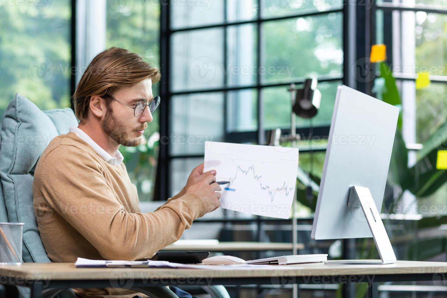 A young man sitting at a desk in the office in front of a computer and talking via call, conducting business training, online meeting. Holds and shows graphics, documents, reports to the camera. photo