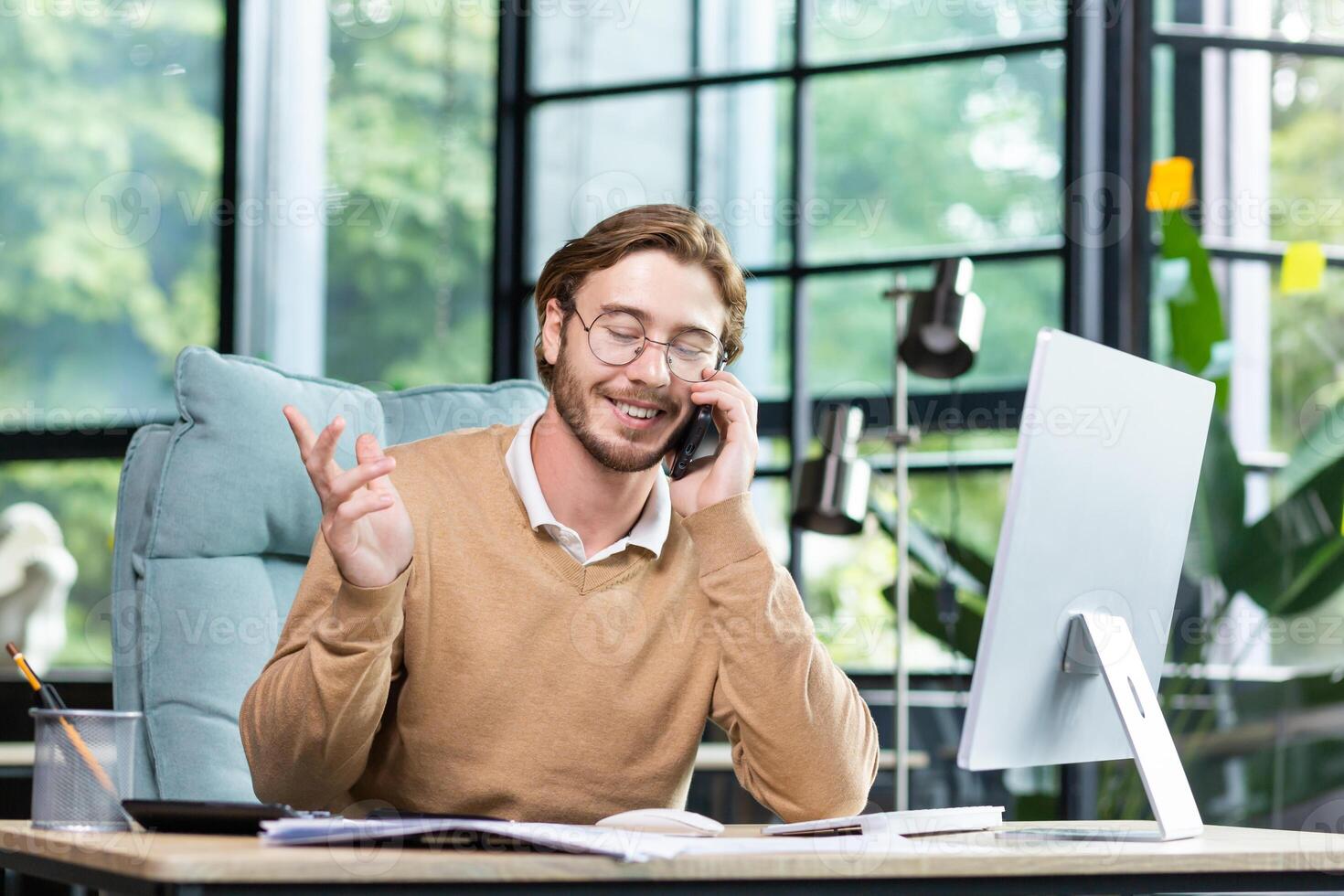 Happy man in the office talking on the phone, sitting at the table in front of the computer, having a successful business conversation, gesturing with his hands. photo