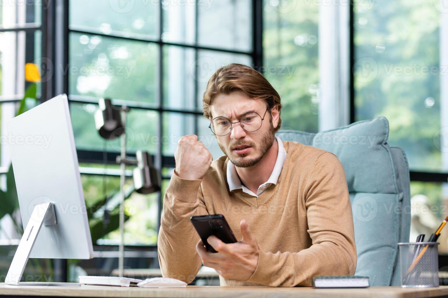 A young man businessman sits in the office at the table and solves questions, problems on the phone. Worriedly looks at the phone, worries, reads messages, checks bills. photo