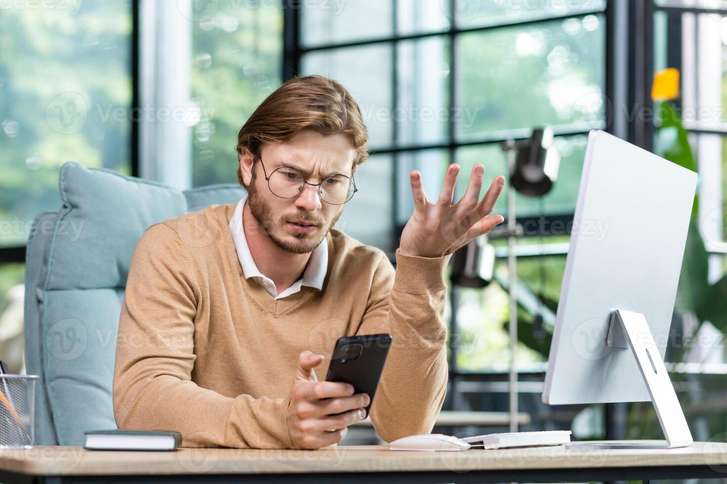 An angry and shocked man sits in the office at the table and looks worriedly at the phone, spreads his hands inexplicably. photo