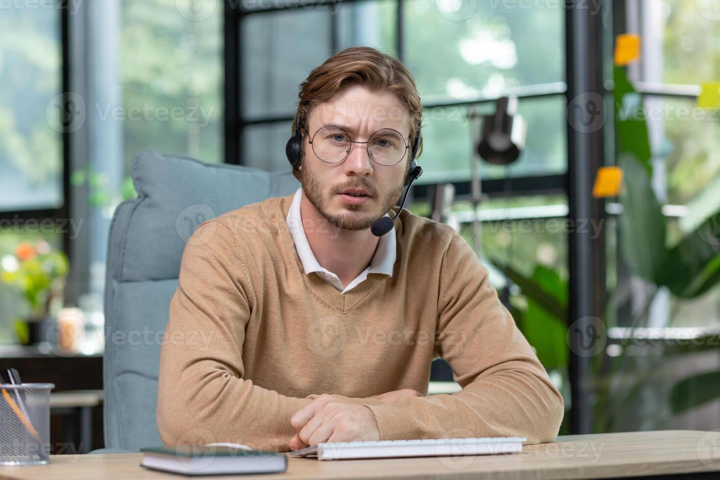 Portrait of a young businessman man in a headset working concentratedly from a home office. Speaks expressively and seriously to the camera, communicates via call. photo