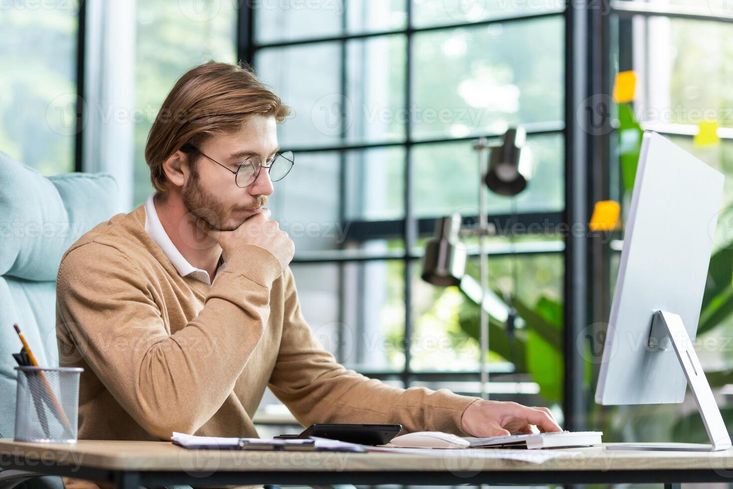 A young businessman sits in the office at a table, works with documents, looks at notes in a notebook, thinks about a strategy, analyzes. He rubs his beard thoughtfully with his hand. photo