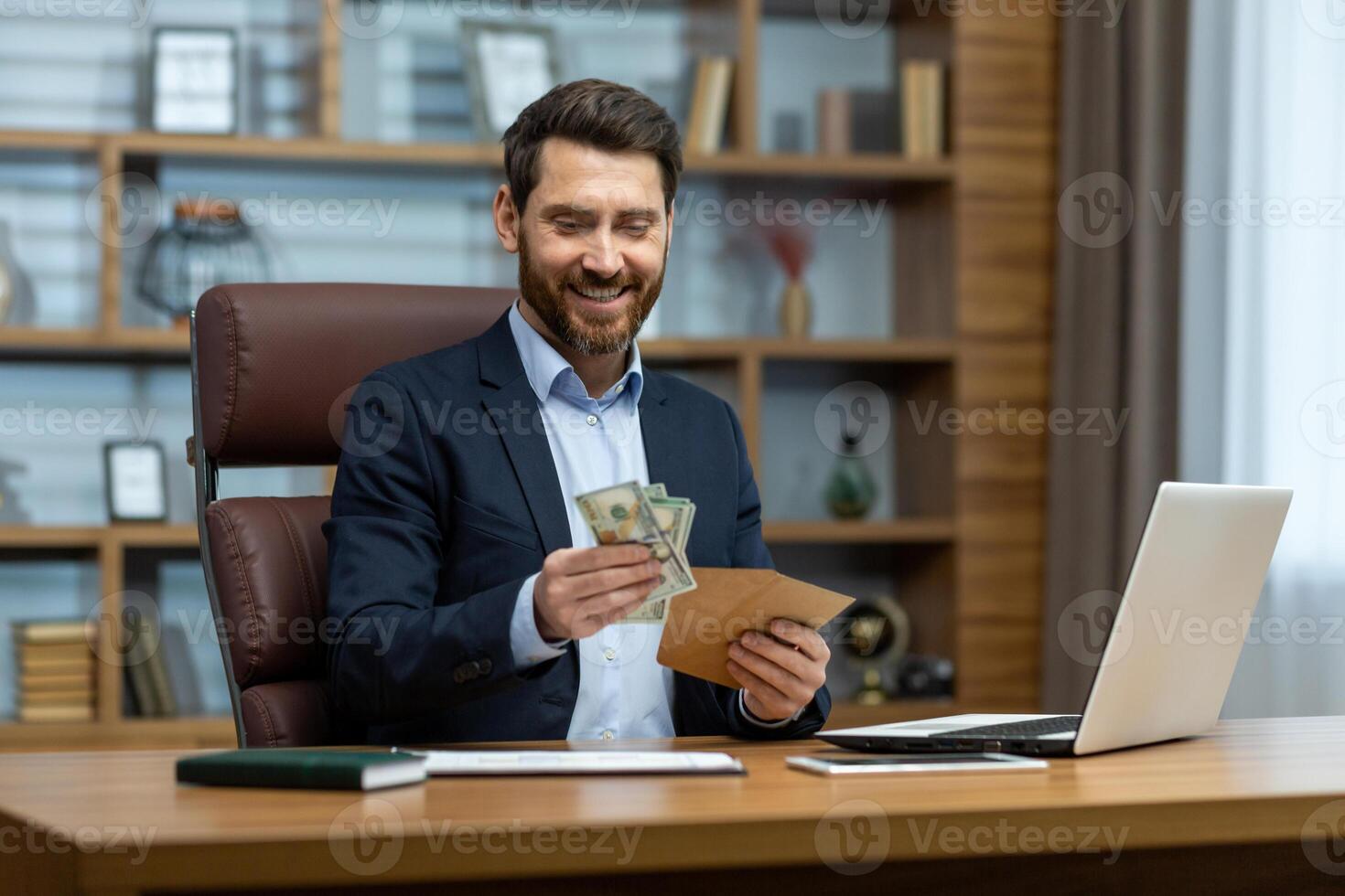 Photo of a happy young businessman sitting at a desk in the office and holding an envelope with cash in his hands, looking satisfied at the received banknotes.