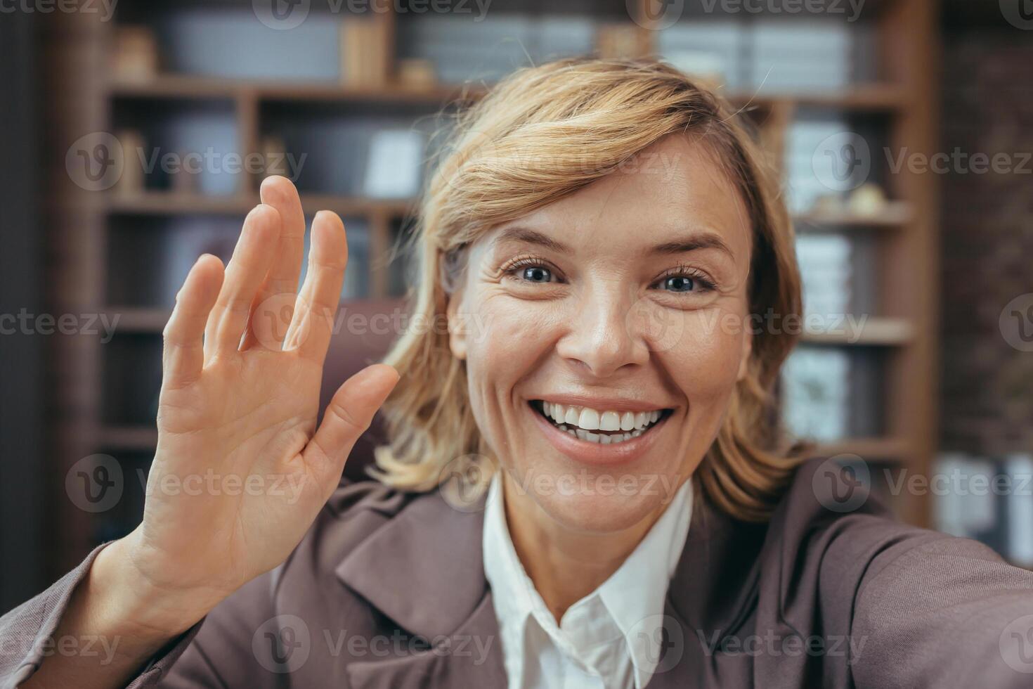 Cheerful mature woman with blonde hair waving hello from her cozy home office, embodying professionalism and friendliness. photo