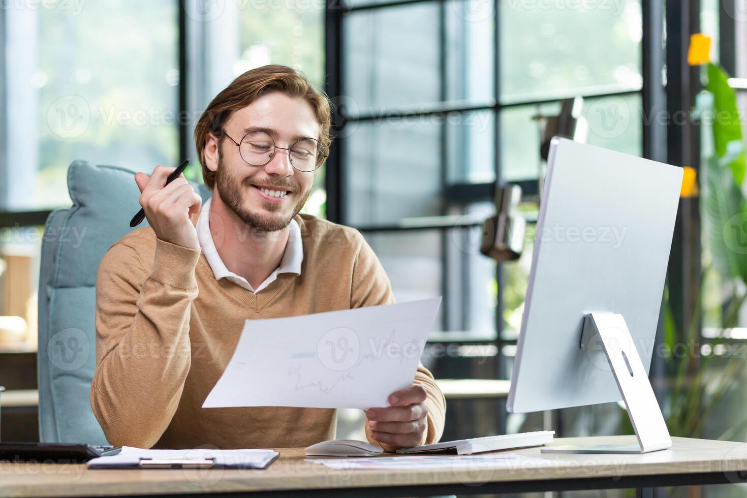 Satisfied young guy doing paperwork in the office, sitting at a desk in front of a computer, joyfully going through business papers, smiling photo