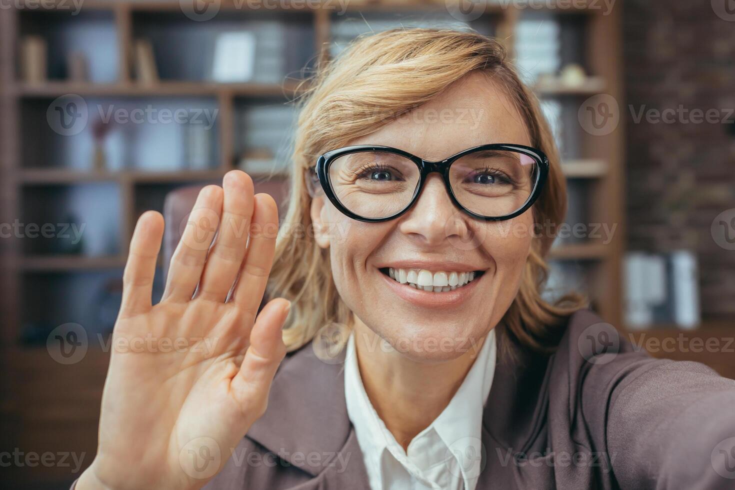 Friendly mature businesswoman with glasses smiling and waving, working remotely from a well-equipped home office setting. photo