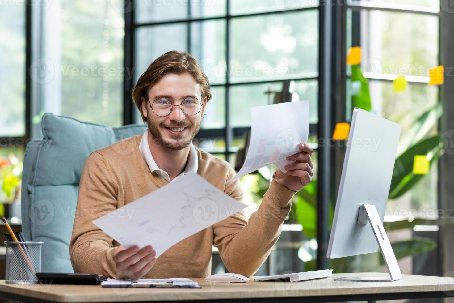 Happy businessman in glasses goes through documents sitting at a table in front of a computer in the office, doing paperwork, smiling and looking at the camera. photo