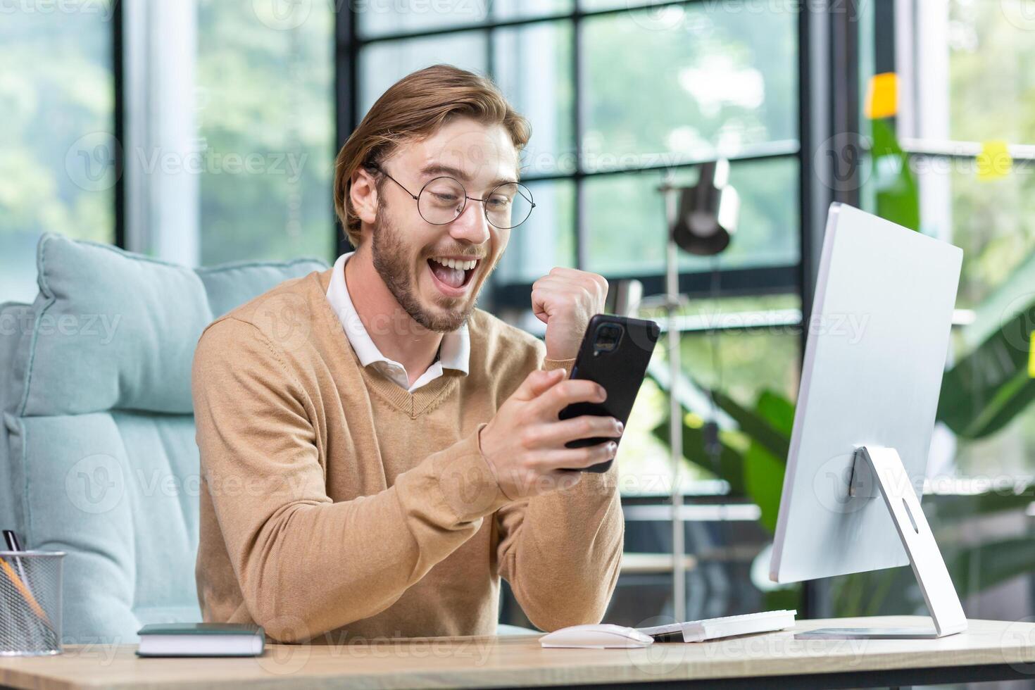 Happy young student in glasses sitting in office at home in front of computer, holding phone in hand, showing victory success gesture with hand, celebrating, smiling. photo
