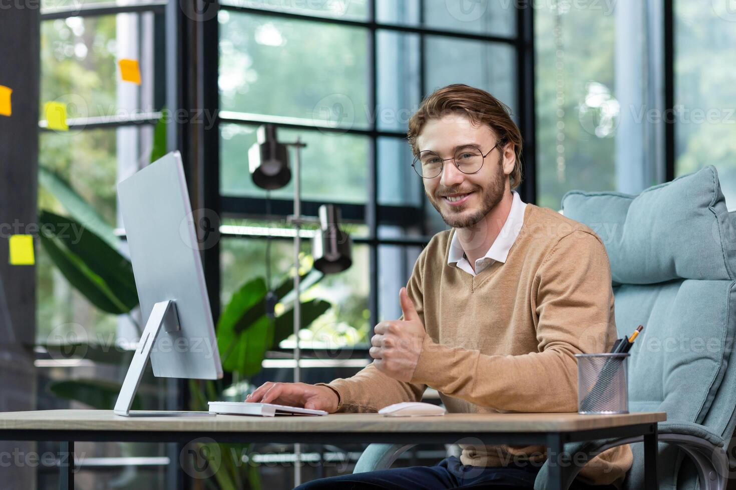 A young male businessman is concentrating on working from a home office at a computer. Smiles at the camera, points a super finger. photo