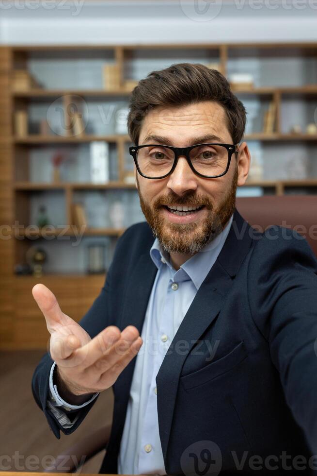 Vertical close-up photo of a young man in a business suit and glasses sitting in the office, talking online and gesturing to the camera of the device.