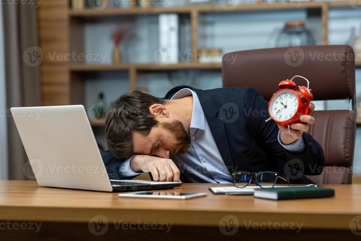 Tired office manager laying down on folded hand on table with personal gadgets and holding red alarm clock. Overworked man having insomnia or chronic fatigue syndrome and taking nap during busy day. photo