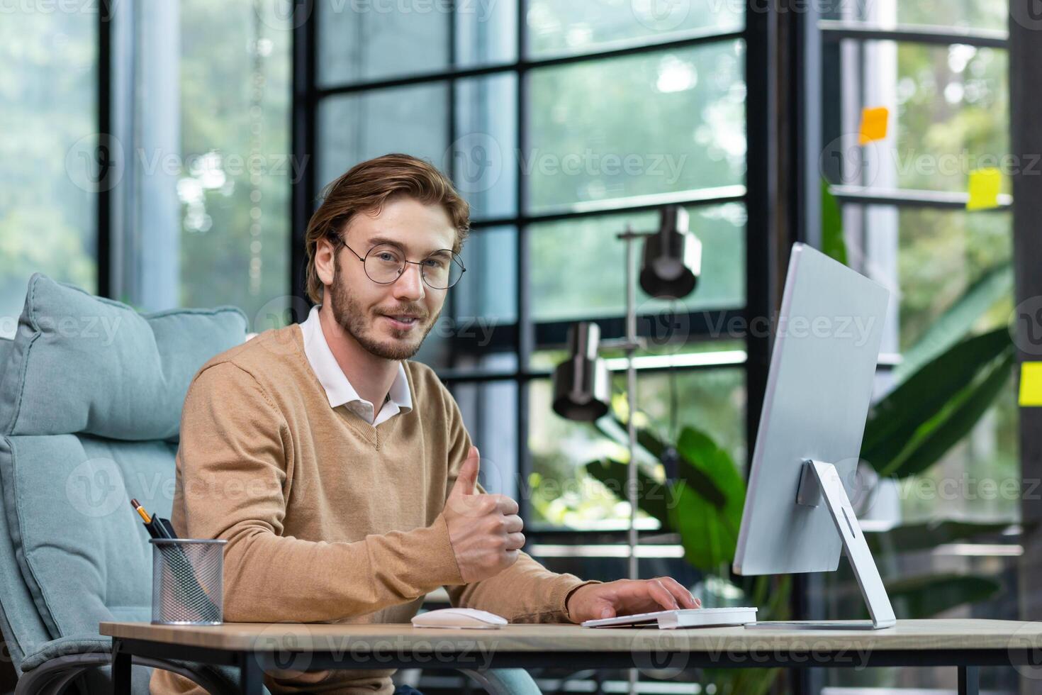 Portrait of a young man in glasses working at a computer in a modern office. He looks at the camera, smiles, shows a successful gesture with a super finger. photo