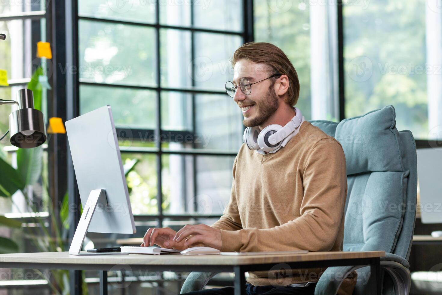Young smiling male student in headphones sits on campus at a desk and studies online at a computer. Writes coursework and diploma work. photo