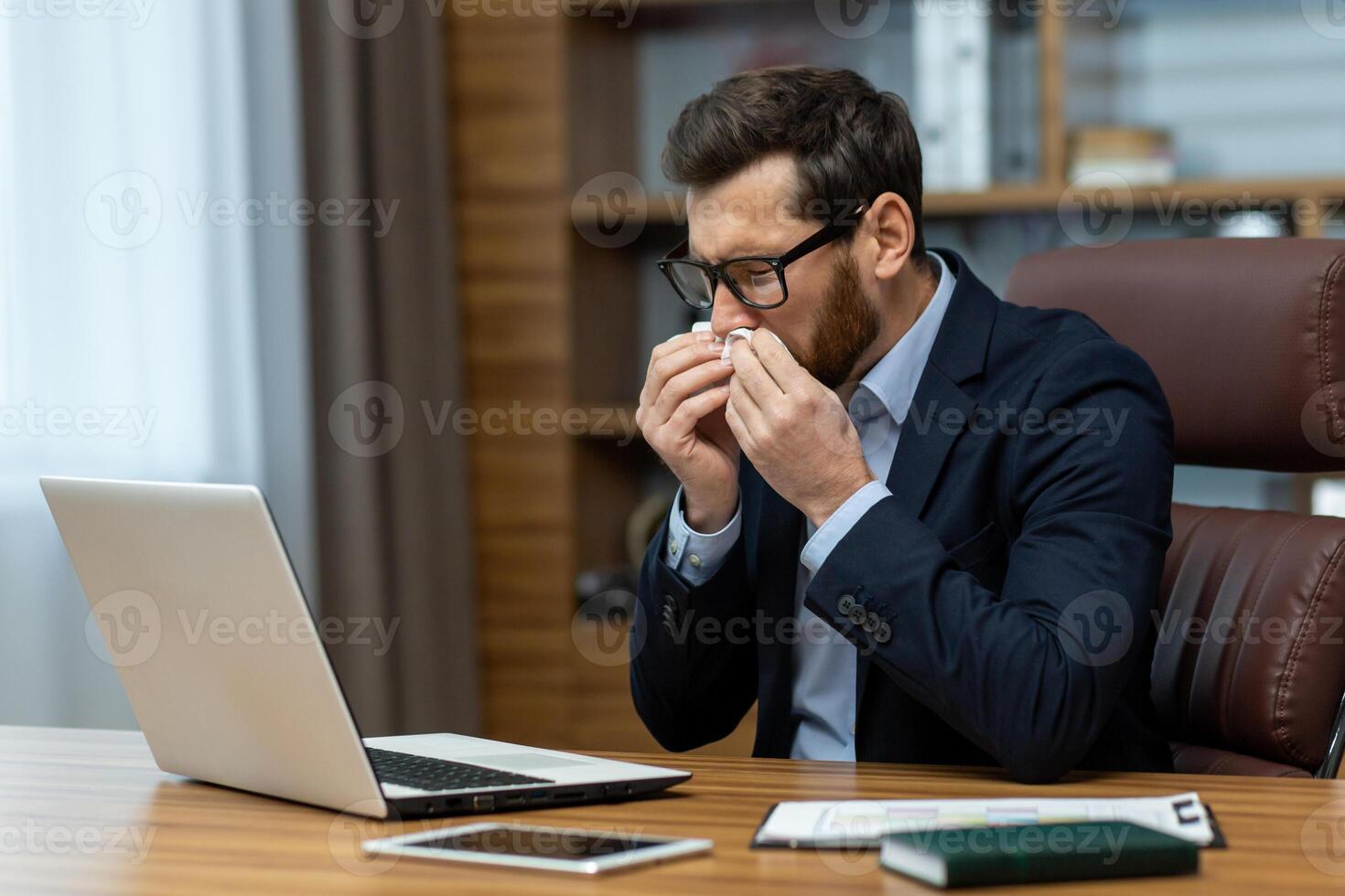 Sick businessman in glasses sitting by desktop with computer and sneezing into paper napkin. Adult man having seasonal allergy or respiratory disease and wiping runny nose with tissue at office. photo
