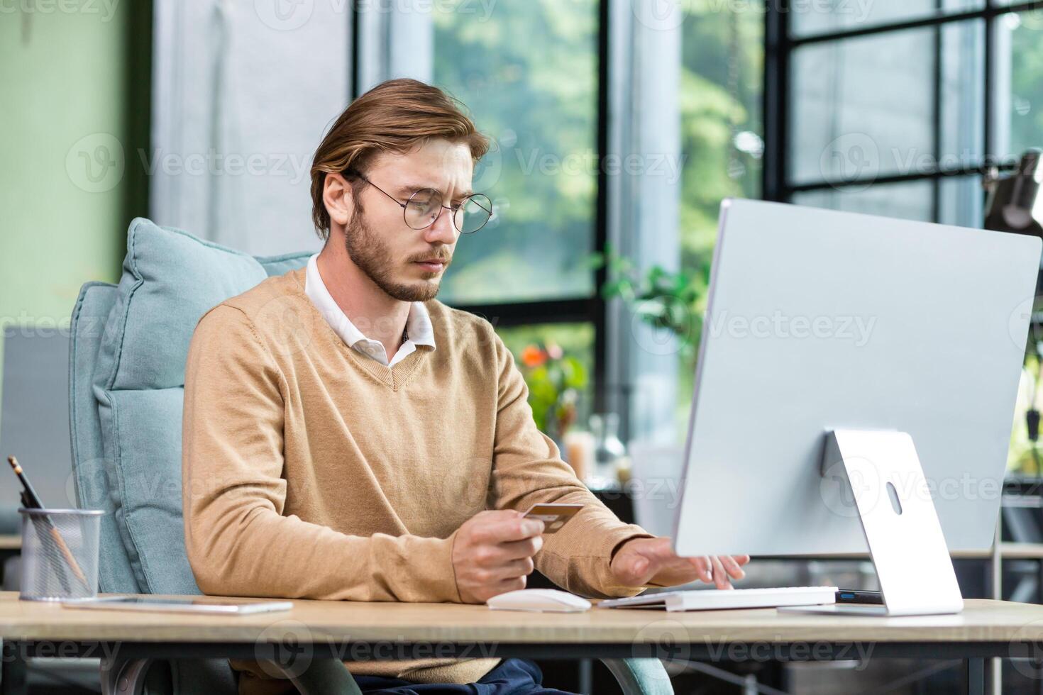 Concentrated young man holds a card in his hands, sits at a desk in the office in front of a computer, makes an online payment, purchases online. photo