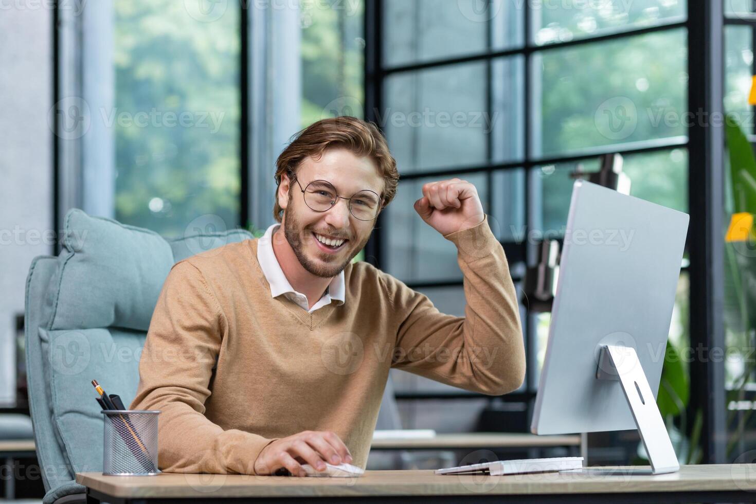 A happy young male student sits in the office, at home in front of the computer, shows a winning gesture of success with his hand, celebrates, looks at the camera smiling. photo