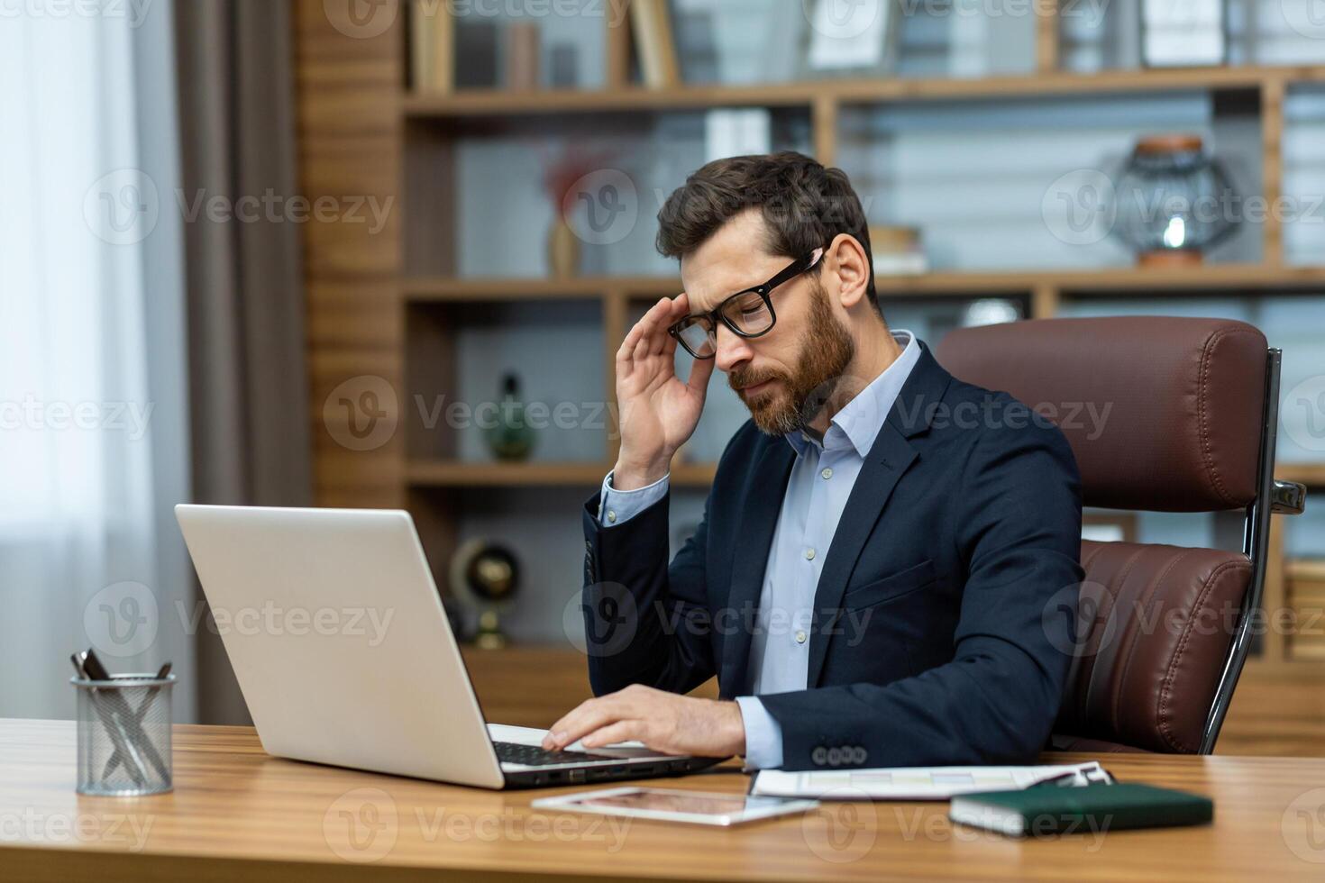 Frustrated administrator experiencing headache while sitting by desk with portable computer in modern interior. Exhausted man feeling pain in temple and getting distracted from working process. photo
