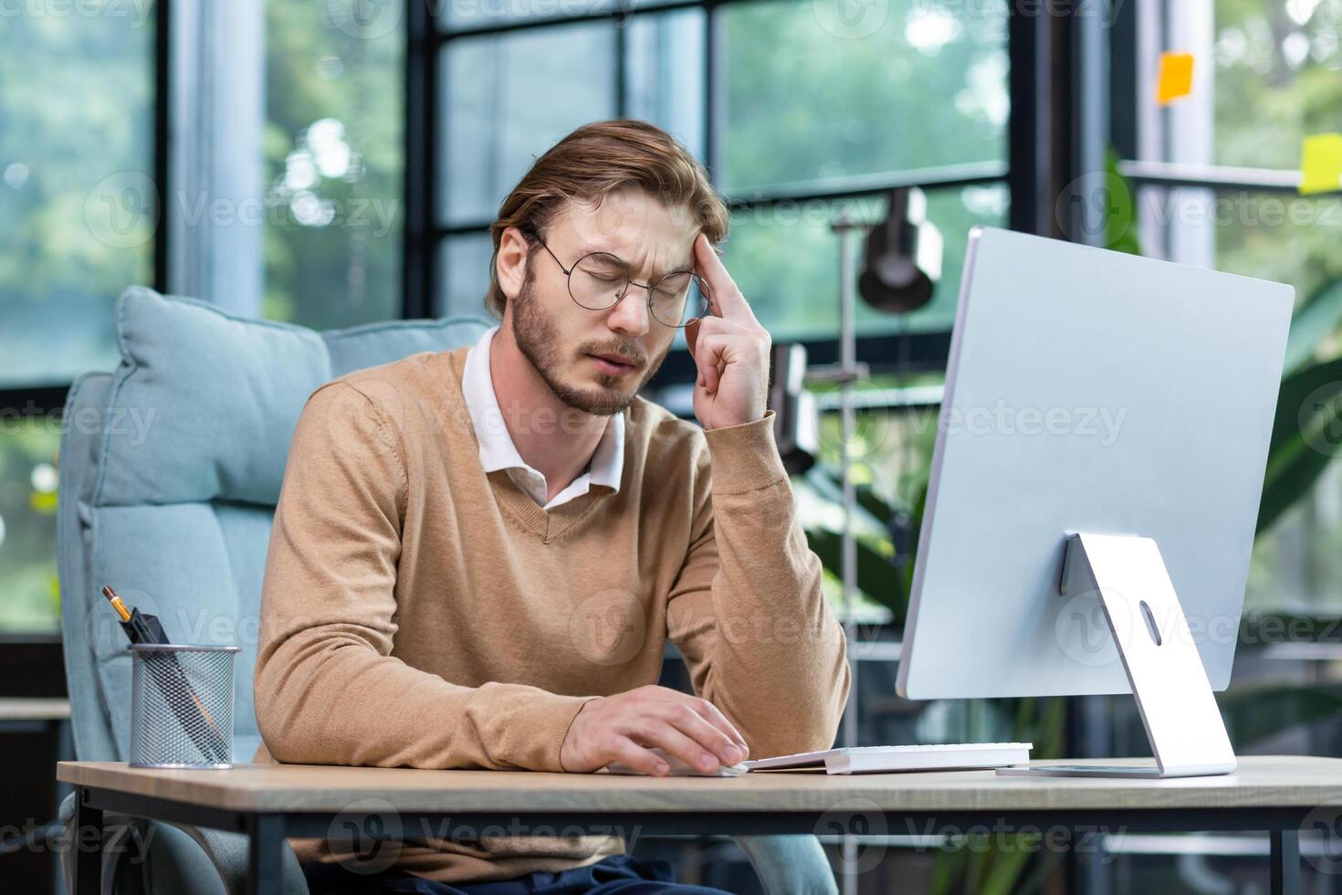 Overtime, deadline. Tired young male programmer, freelancer sits in the office and tries to work at the laptop. Bored, he sits at the table and rests his head on his hand. photo