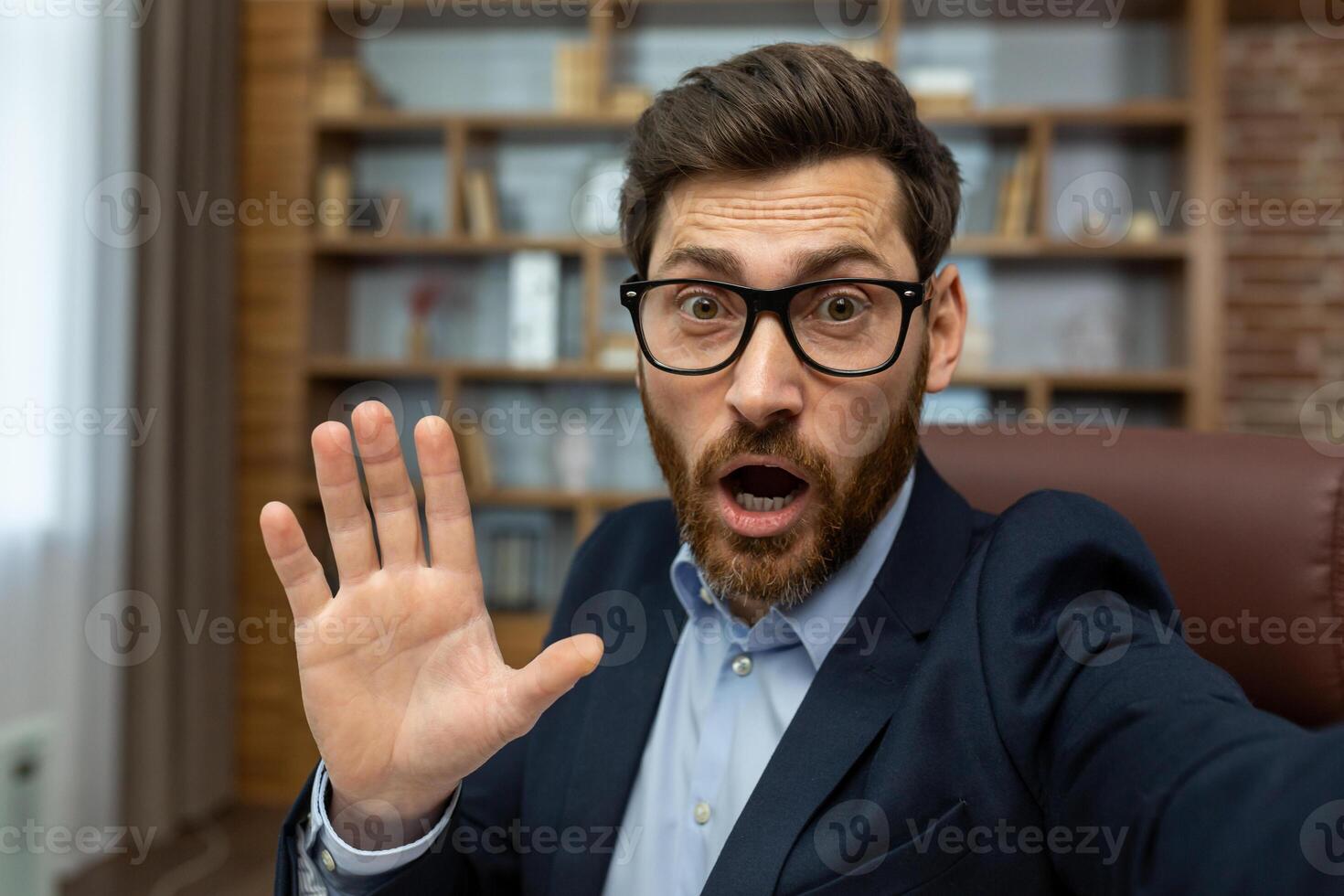 Mature man in a business suit appears shocked or overwhelmed, gesturing with his hand as if saying no or stop while in a messy home office setting. photo