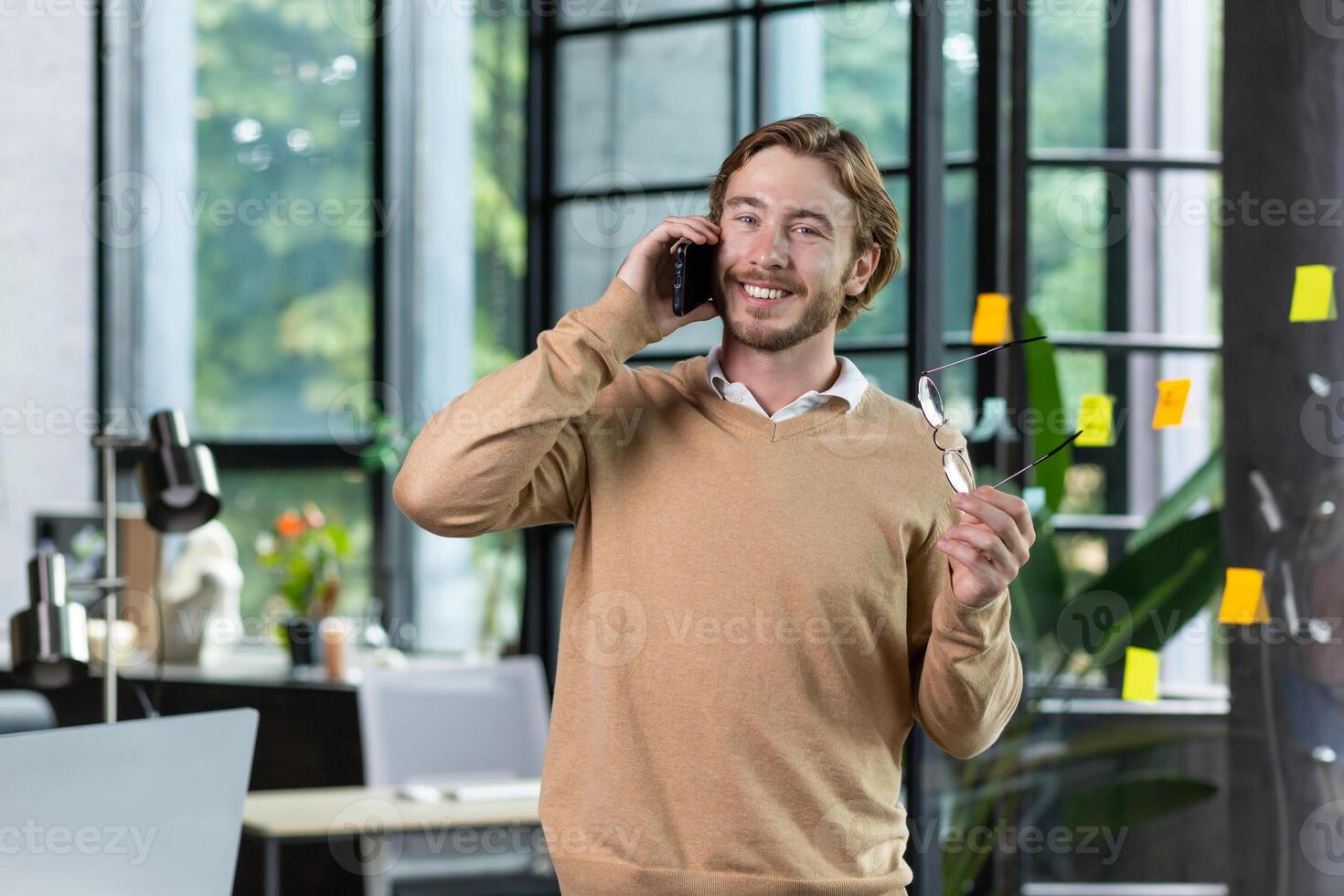 A young freelancer man is standing in the office and talking on the phone. He holds glasses in his hands, looks at the camera smiling. photo