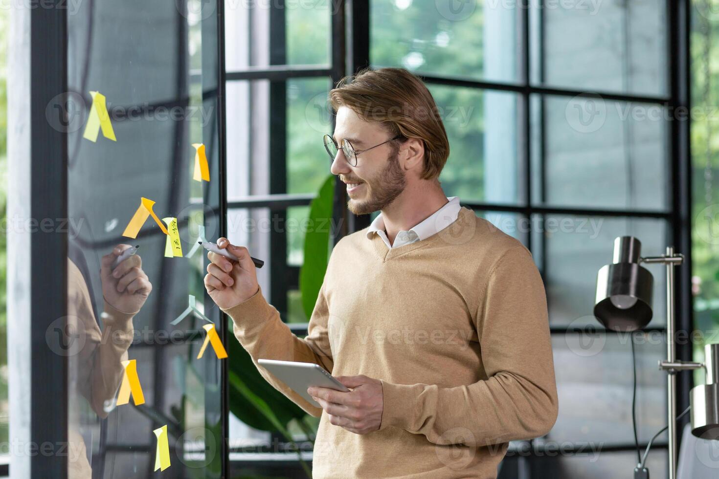 Happy man in casual clothes standing near a glass note board and sticking colorful stickers with reminders and plans on it, writing something with a pen, office work planning concept. photo