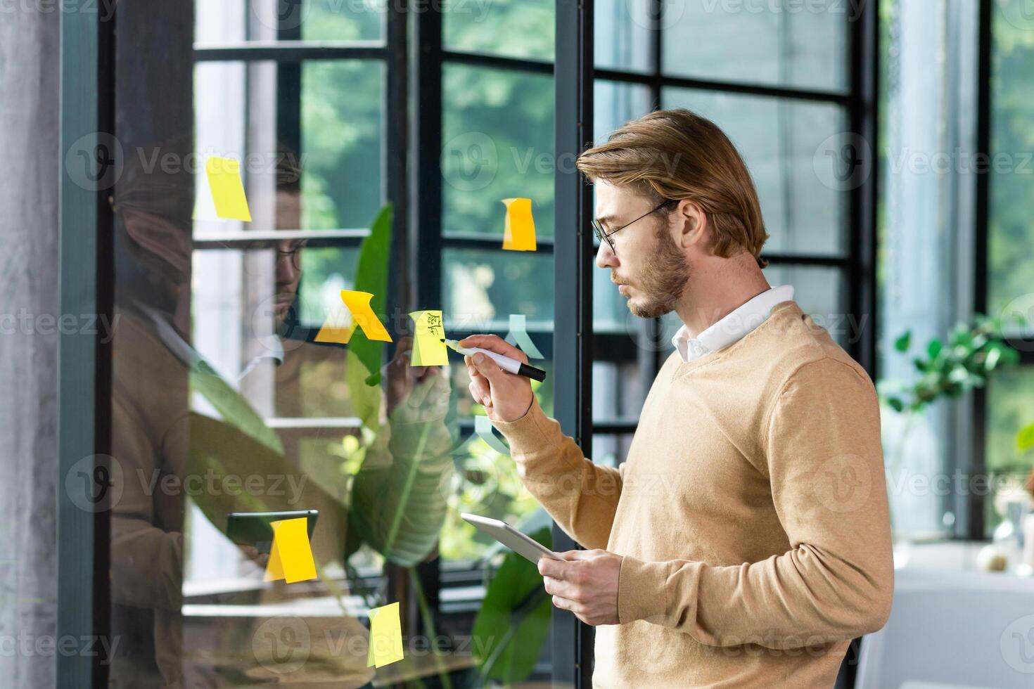 A young man in casual clothes stands near a transparent note board and attaches stickers with reminders and plans to it, the concept of work planning in the office. photo