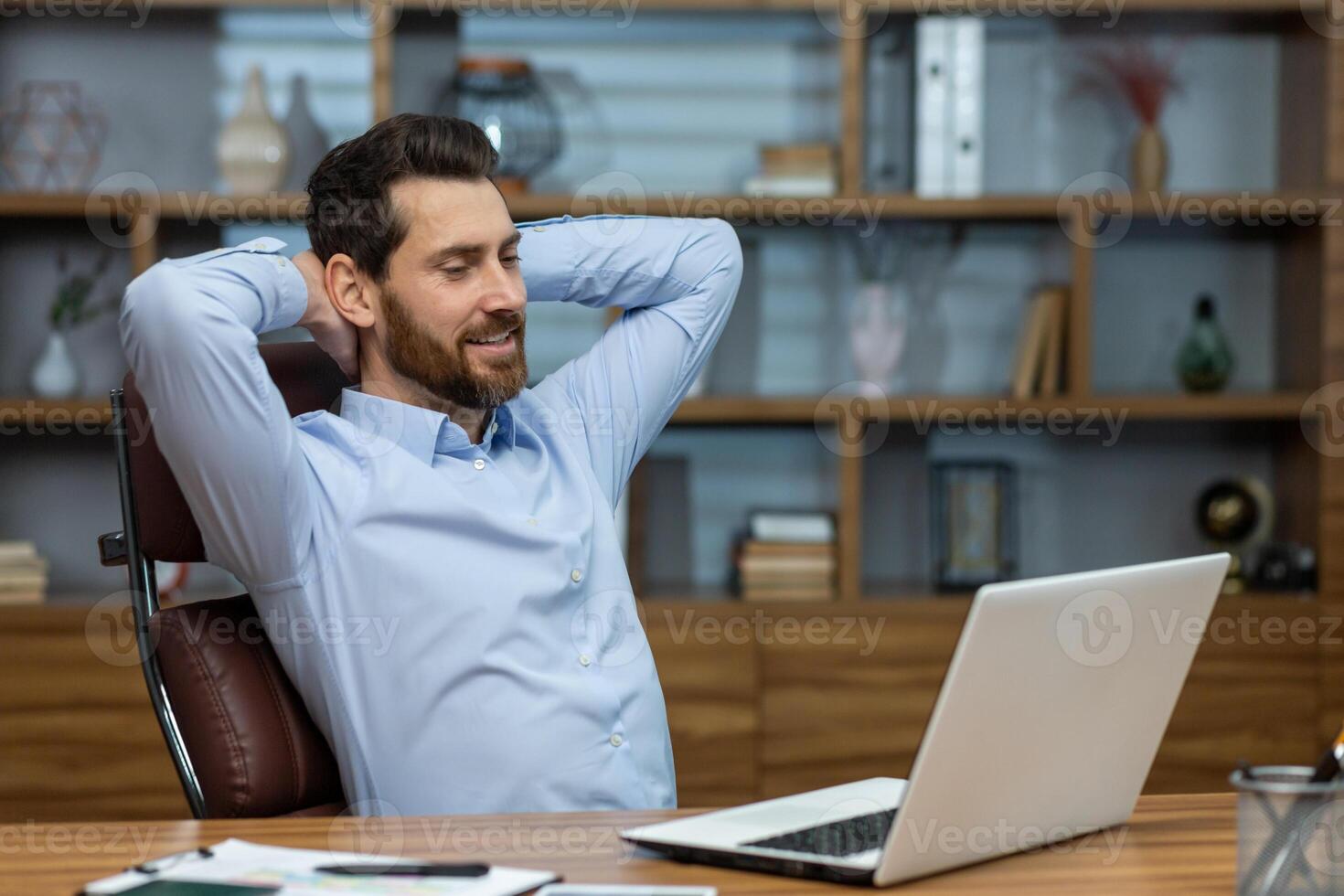 Relaxed brunette man leaning back and holding hands behind head in personal workspace with pc. Calm head of company taking break during busy day and enjoying daydreaming in comfortable chair. photo