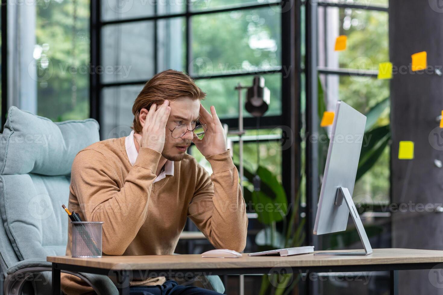A young male programmer, IT developer codes, thinks about a new project. He is sitting concentrated at the desk in the office, holding his head thoughtfully with his hands, looking at the monitor. photo