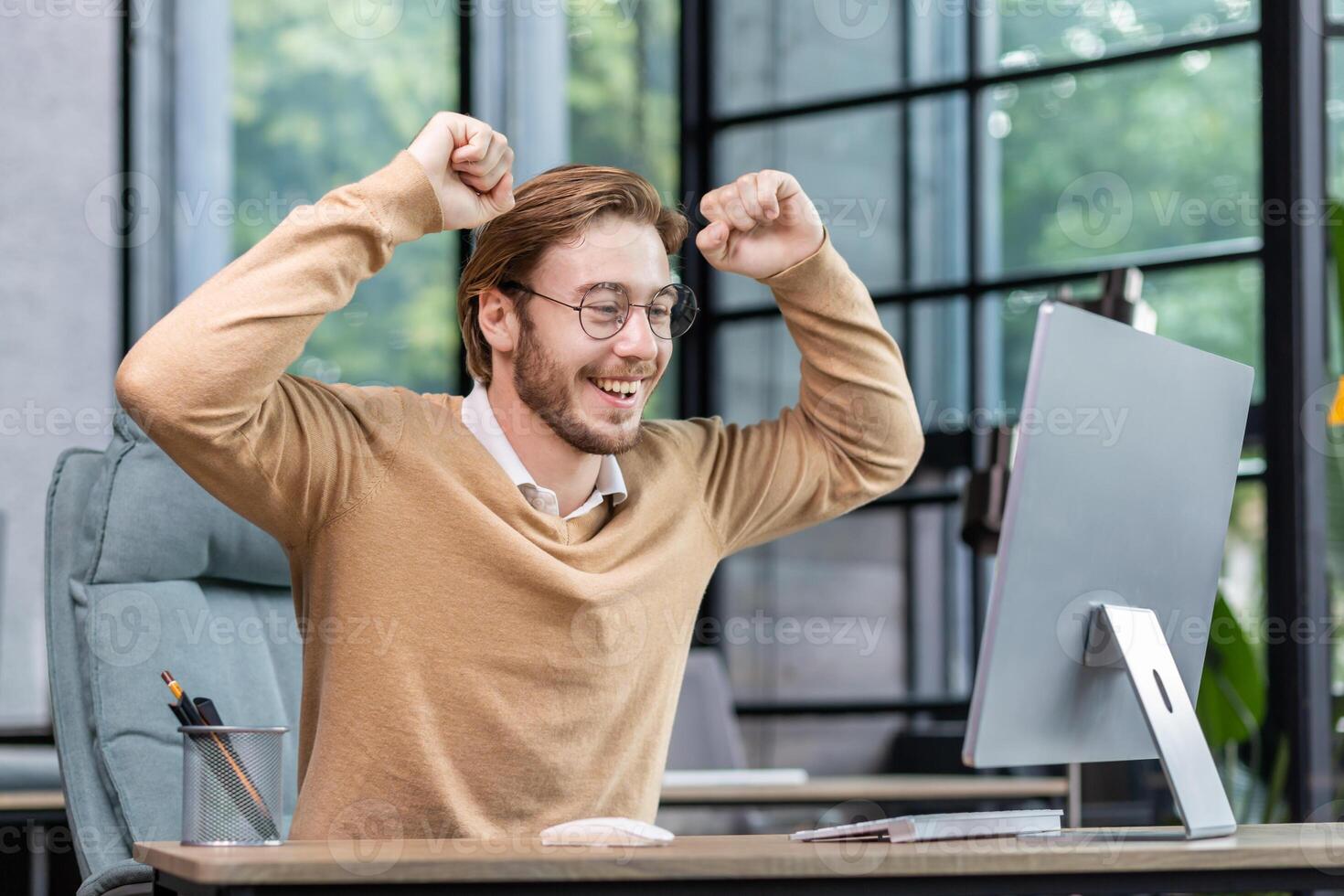 Smiling young male programmer, freelancer sits at the table, looks happy at the computer monitor. Raised his hands up, rejoices in success, victory. photo
