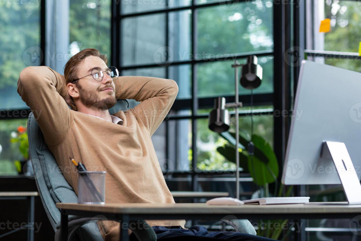A young male student is taking a break from studying after an online exam. He is sitting in the office at a table with a computer, hands behind his head, leaning back in a chair, smiling. photo