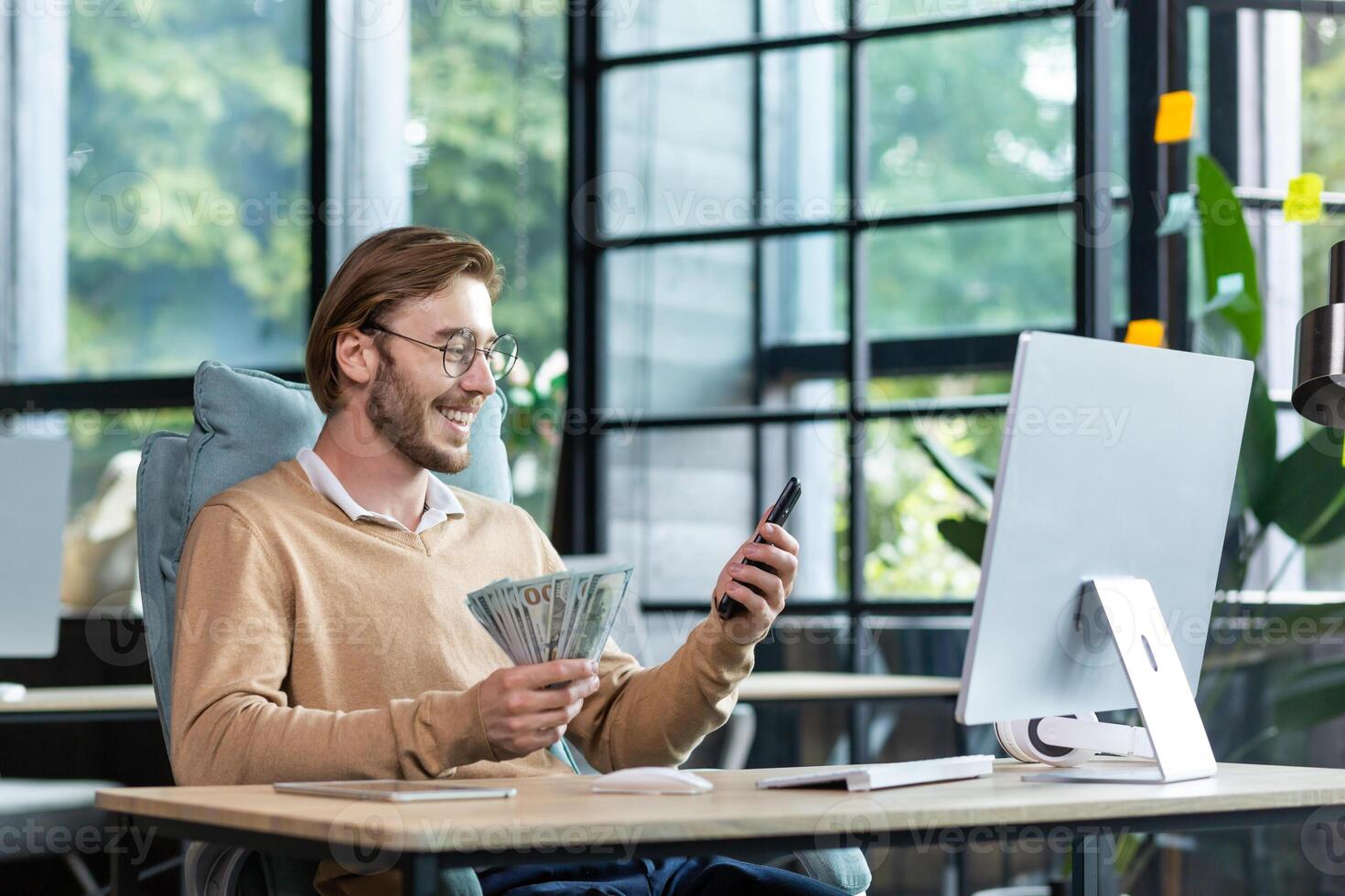 A happy young man is sitting at the table in the lobby, working at the computer. Holds cash, dollars and uses the phone. photo