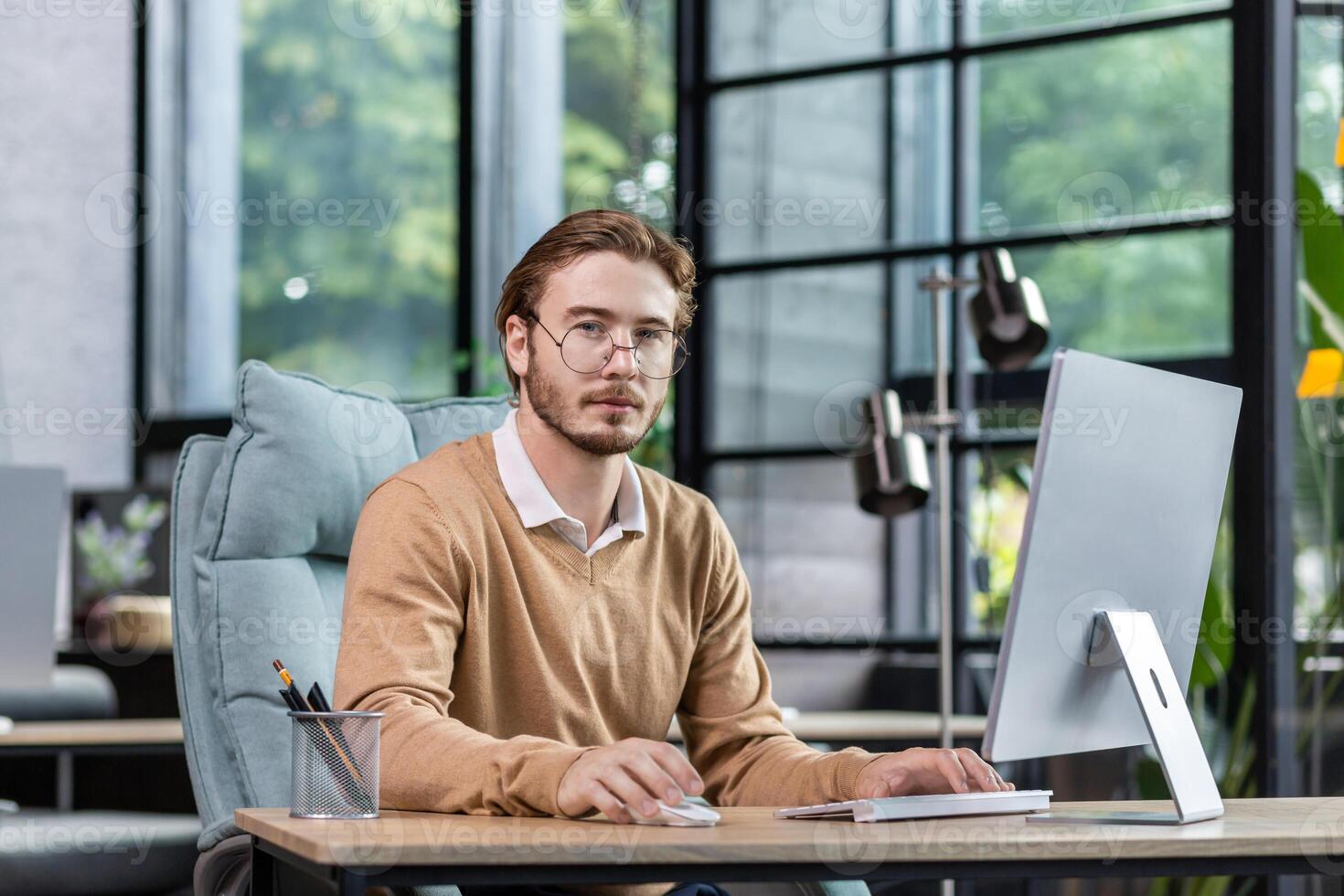 Portrait of a young man, psychologist, psychotherapist sitting in the office, working at the computer, consulting online, talking via call with patients. He looks seriously into the camera. photo