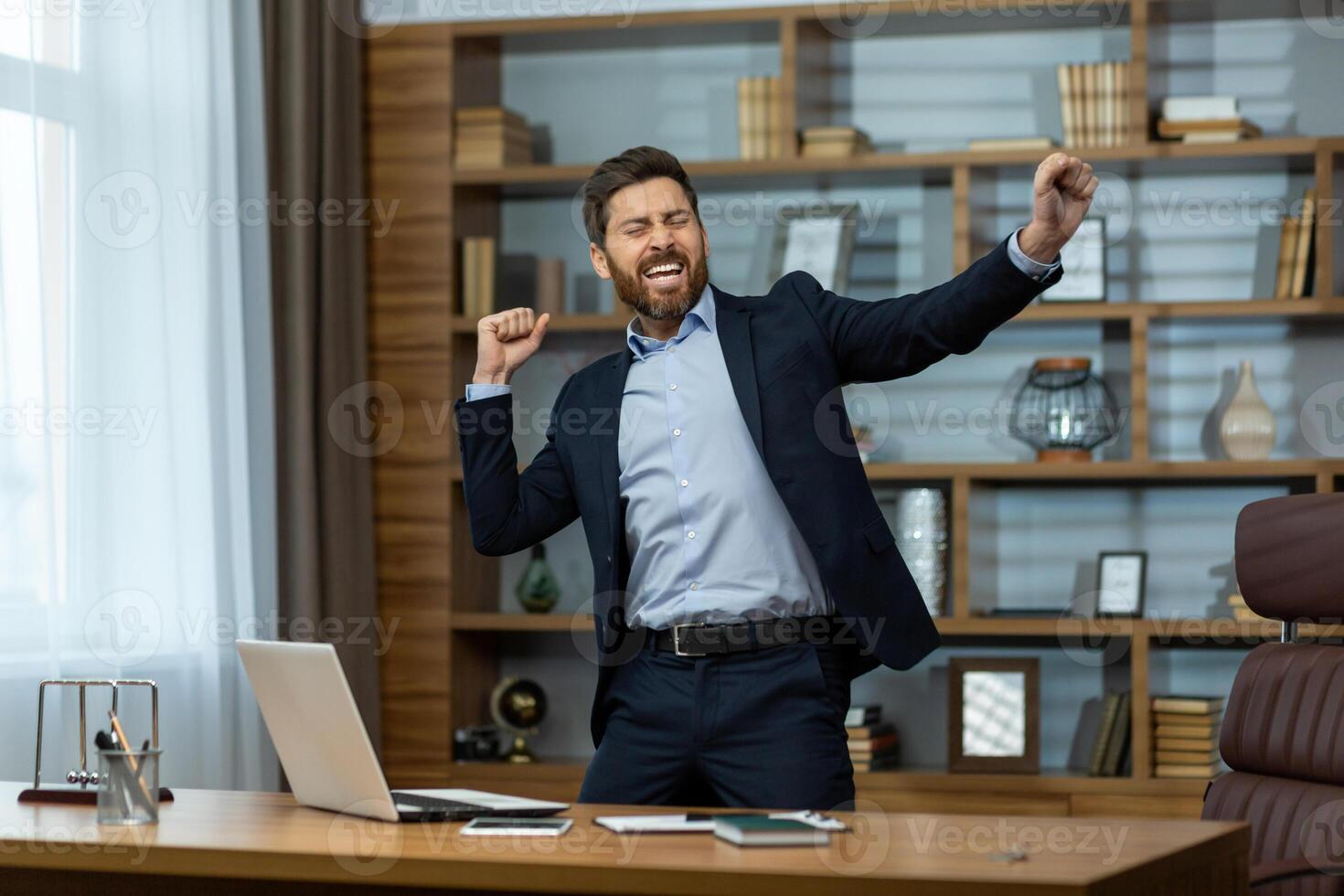 A happy young man in a business suit standing in the office at the desk with a laptop and dancing with his hands up, rejoicing at the success and result, resting during a break. photo