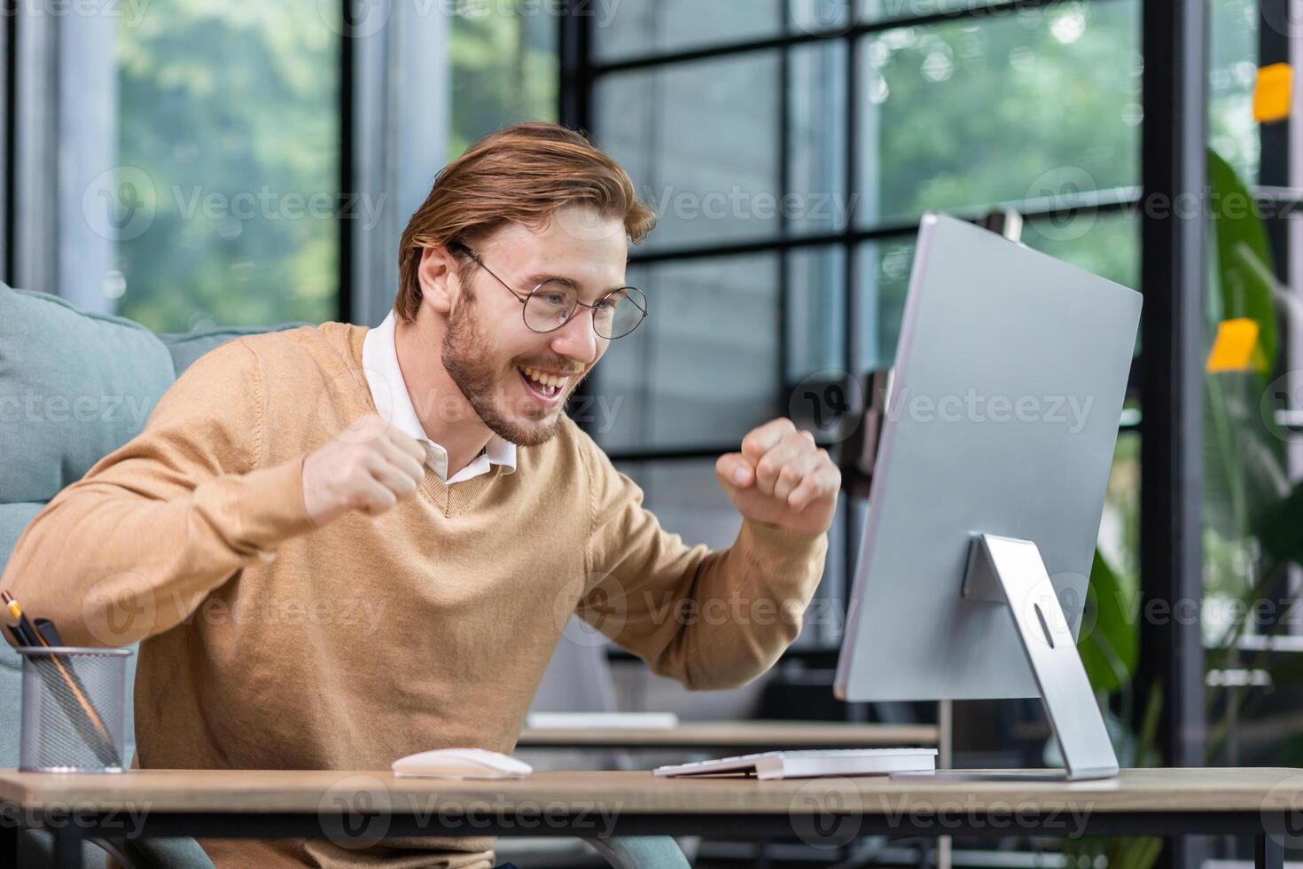 Young man in office celebrating victory and successful achievement results, businessman holding hands in triumph gesture and reading good news from computer , blond programmer in casual clothes. photo