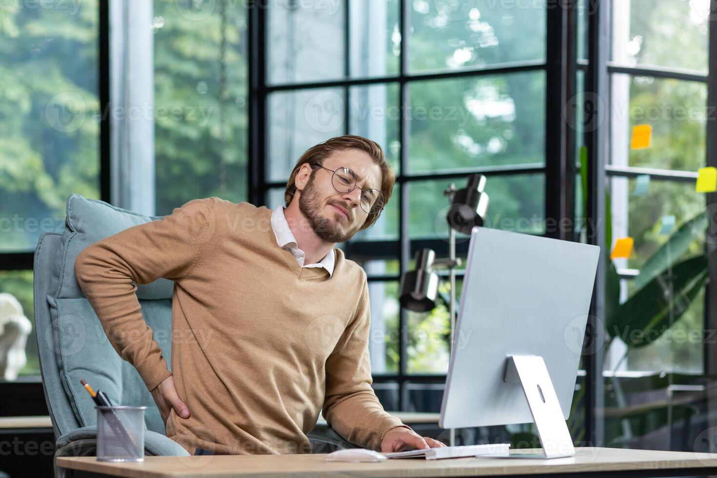 Tired businessman at workplace has severe back and lower back pain, young man working sitting at table with laptop and computer in middle of office. photo