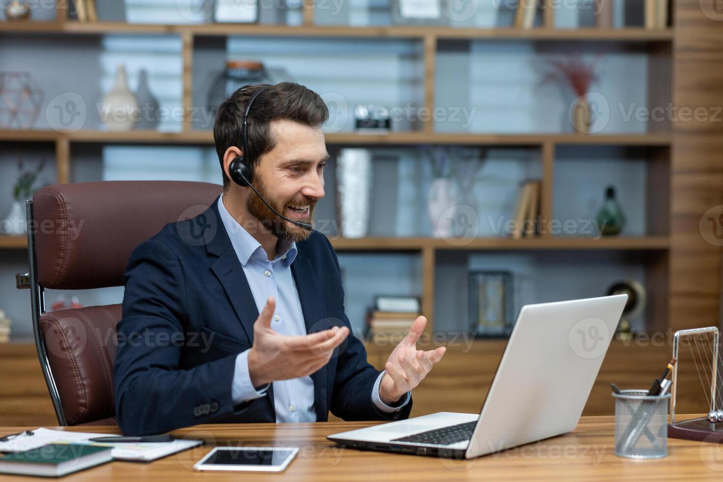 Positive man in wireless headset with micro hosting conference while sitting by desktop with computer. Millennial businessperson using modern technology for online communication with partners. photo