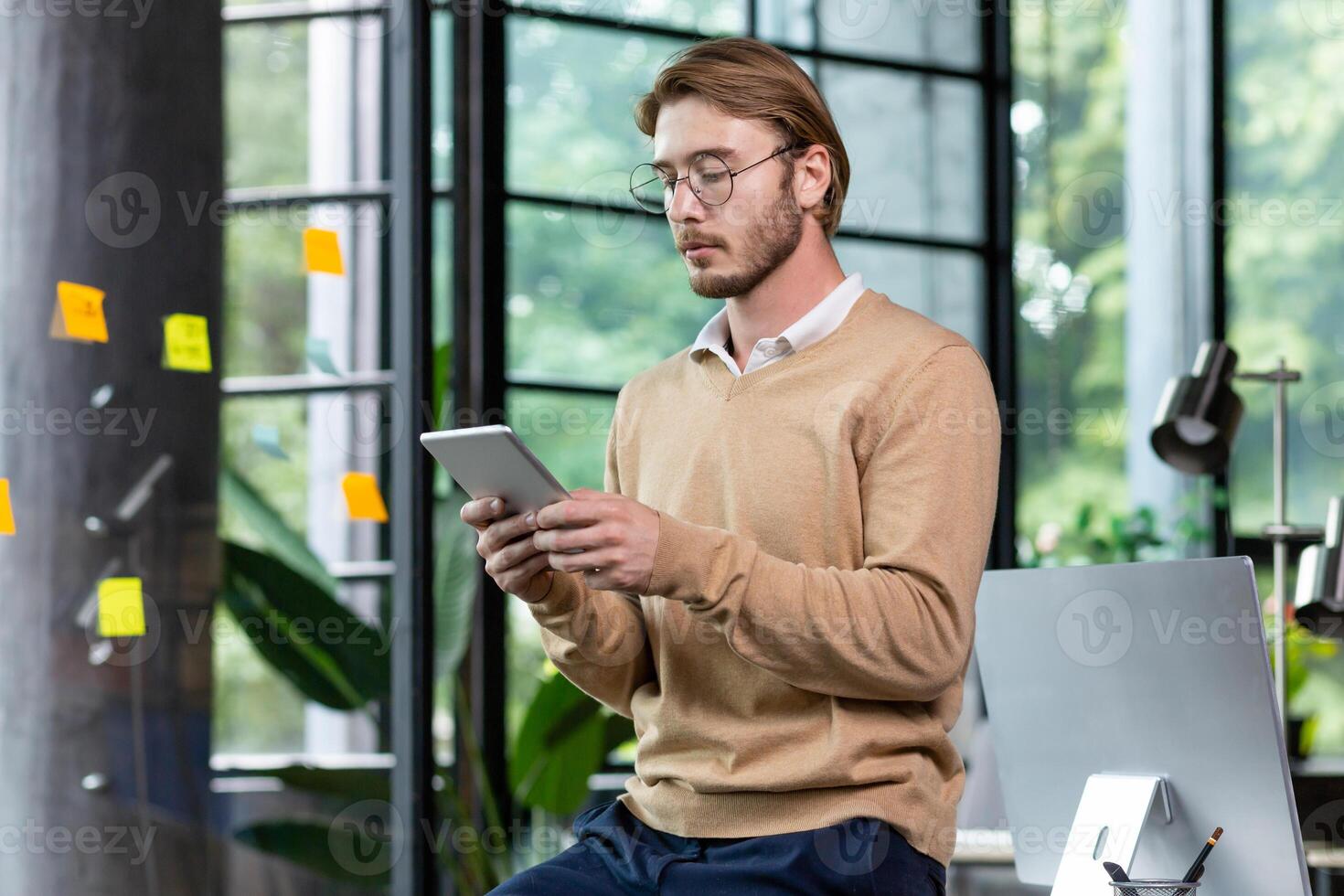Young successful thinking programmer inside loft style office uses tablet computer, young blond man in casual clothes reads news online, serious businessman browses internet pages. photo
