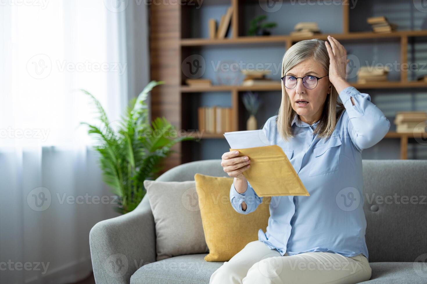 Shocked elderly woman receiving unexpected news while reading letter on sofa at home. Senior lady with glasses and hand on head expresses surprise or worry. photo