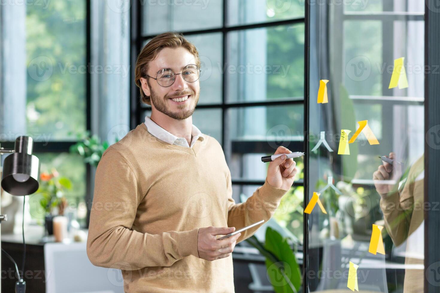 A young designer, programmer, freelancer man is standing in the office, holding a tablet, drawing a progress graph on a transparent board. Glue colored, stickers, hints. He looks at the camera, smiles. photo