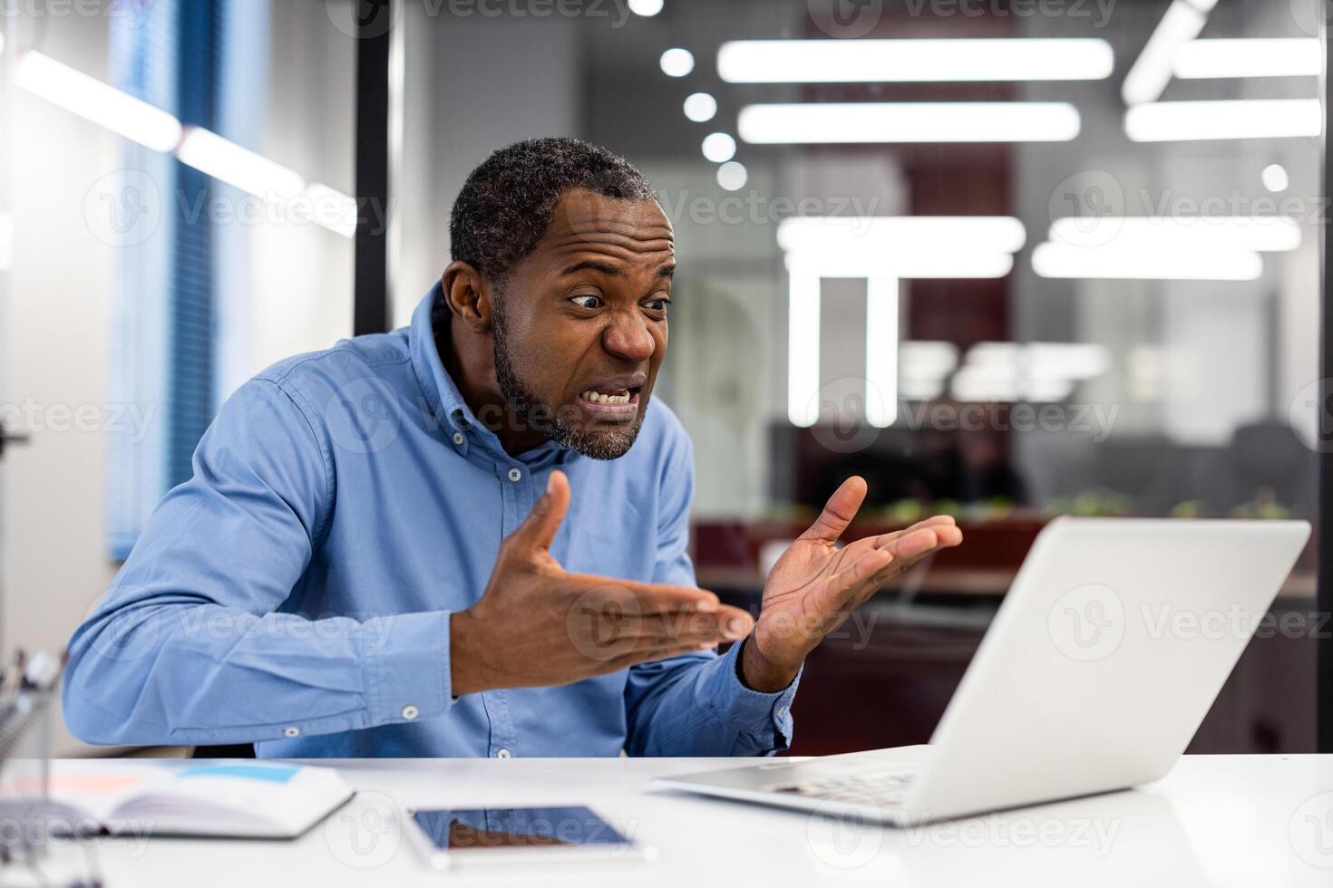 Frustrated businessman sitting in office expressing stress and confusion towards laptop screen. Business setting with modern interior, and desk items depicting workplace challenges and emotions. photo