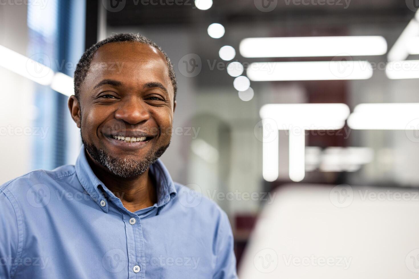 Business professional in blue shirt smiling while working at modern office desk. Bright lighting in background. Confident and positive attitude representing workplace happiness and productivity. photo