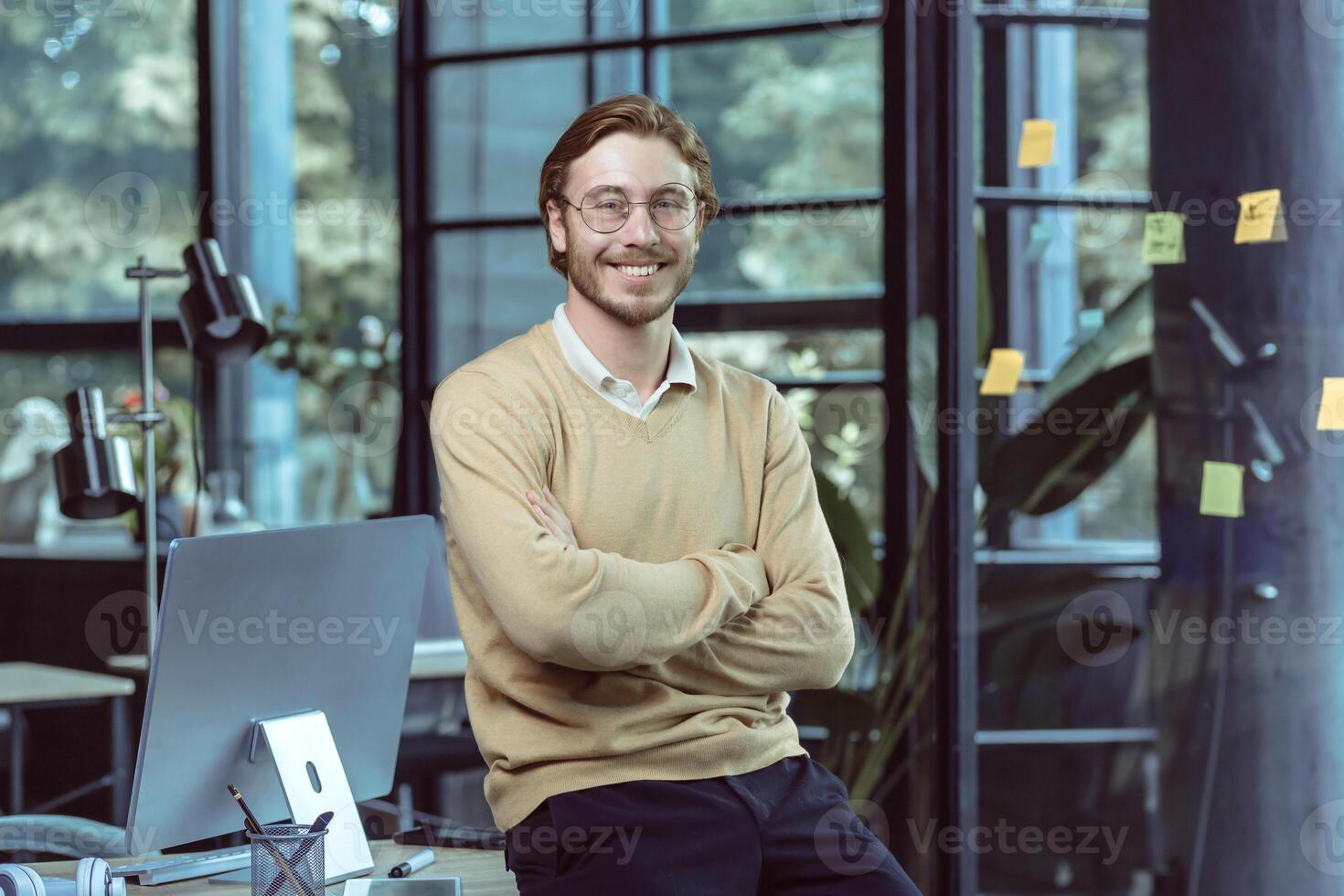 Portrait of successful young IT specialist young engineer programmer in modern loft office smiling and looking camera with crossed arms, businessman business owner wearing glasses and casual clothes photo