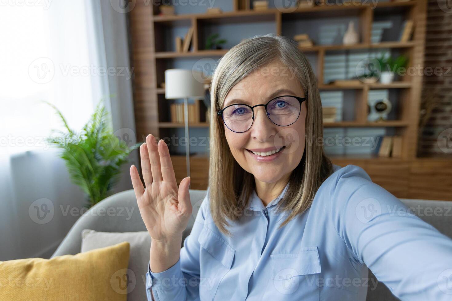 A cheerful older woman with glasses waves and smiles during a call, seated on a cozy couch in her living room. The background displays a modern bookshelf and houseplants photo
