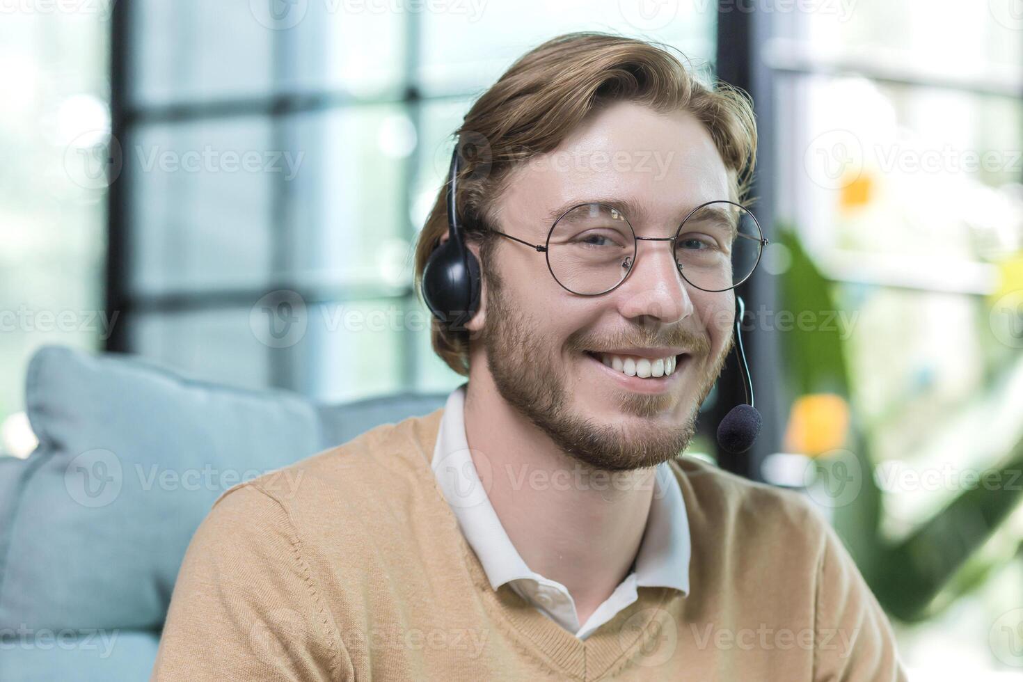 Close-up photo of man wearing headset for call, businessman working inside office building, smiling and looking at camera, employee at work wearing glasses