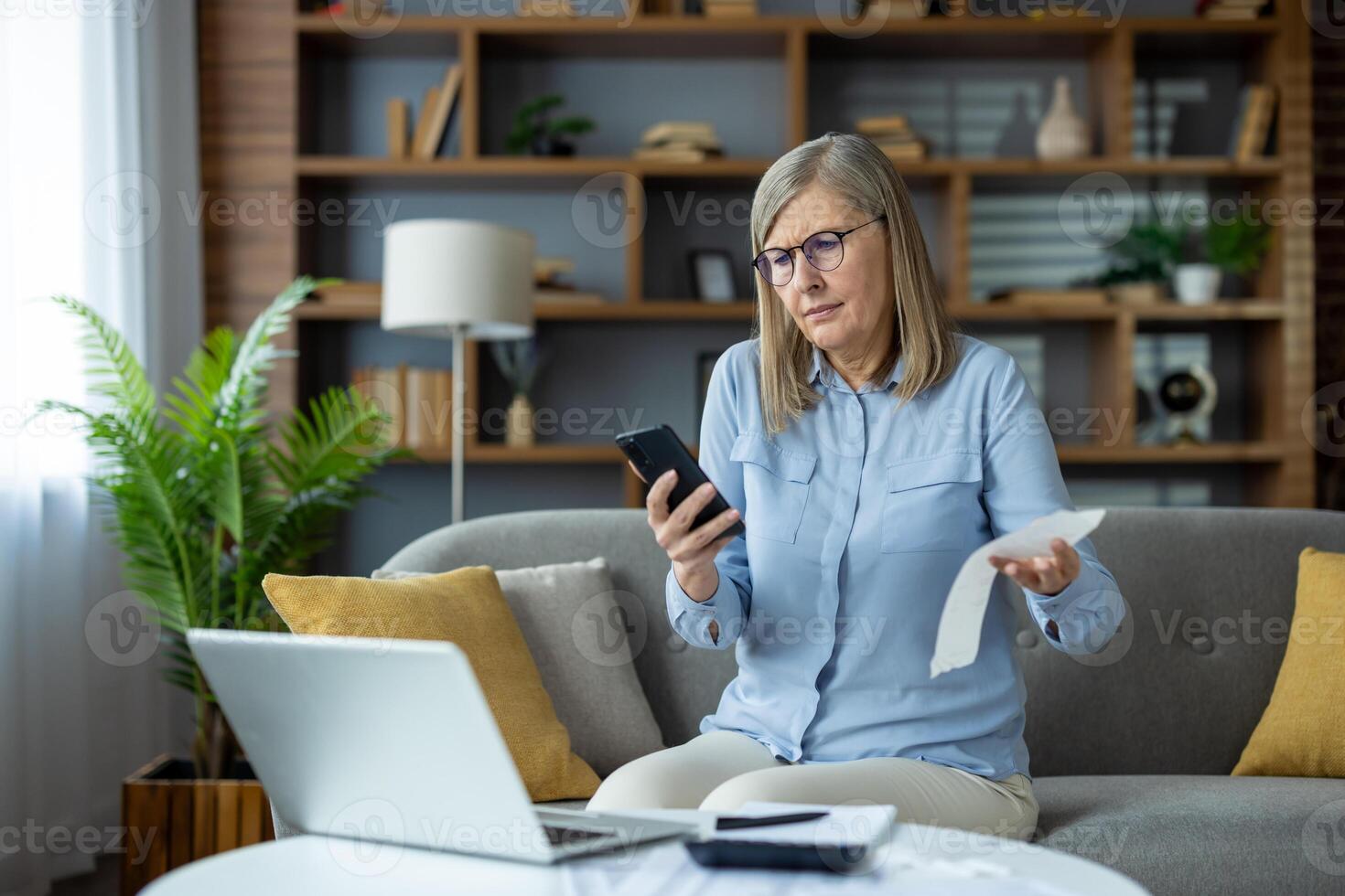 Senior woman sitting on a couch at home, holding a smartphone and a receipt, looking concerned while reviewing her expenses on a laptop. Concept of budgeting, financial planning, photo