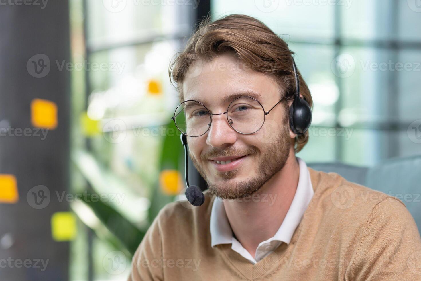 Portrait of young man with headset for call inside office, close-up tech support worker in glasses smiling and looking at camera, businessman blond customer service online help. photo