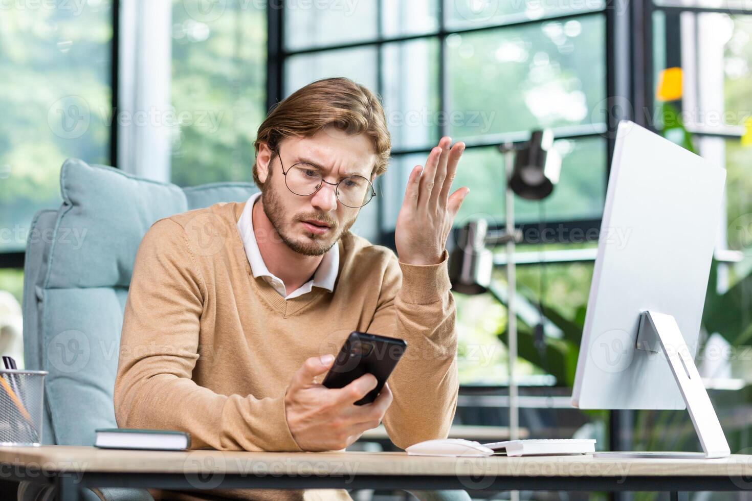 Upset young male programmer, freelancer sitting in the office at the table, holding the phone in his hands, looking worriedly. There was a problem, bad news, messages, checking the account. photo