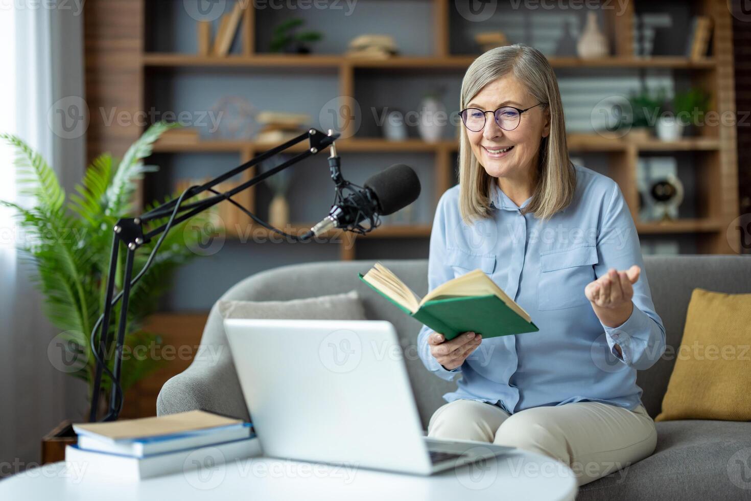 Senior woman recording podcast at home, holding a book and smiling, seated in front of a laptop and microphone. photo