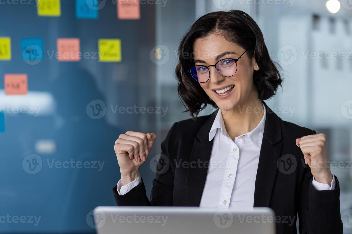 Happy businesswoman in formal attire celebrating a success with fists raised, working on laptop in modern office. photo