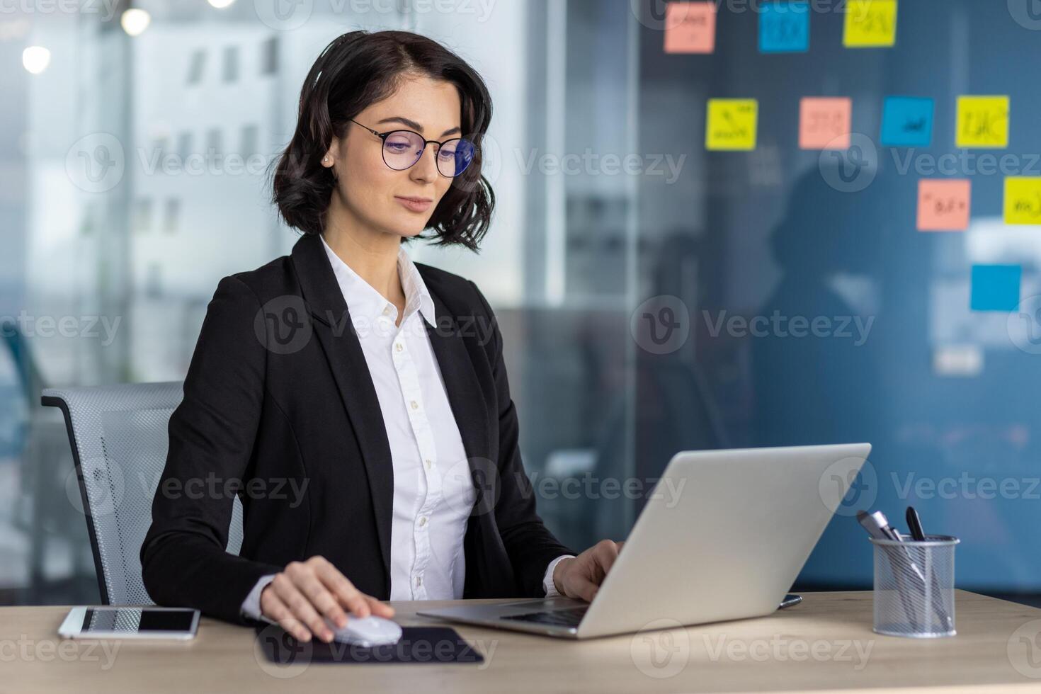 Confident businesswoman in formal attire working on laptop in modern office. Professional setting with sticky notes on glass wall, tablet and pen holder on desk. Concept of corporate work photo
